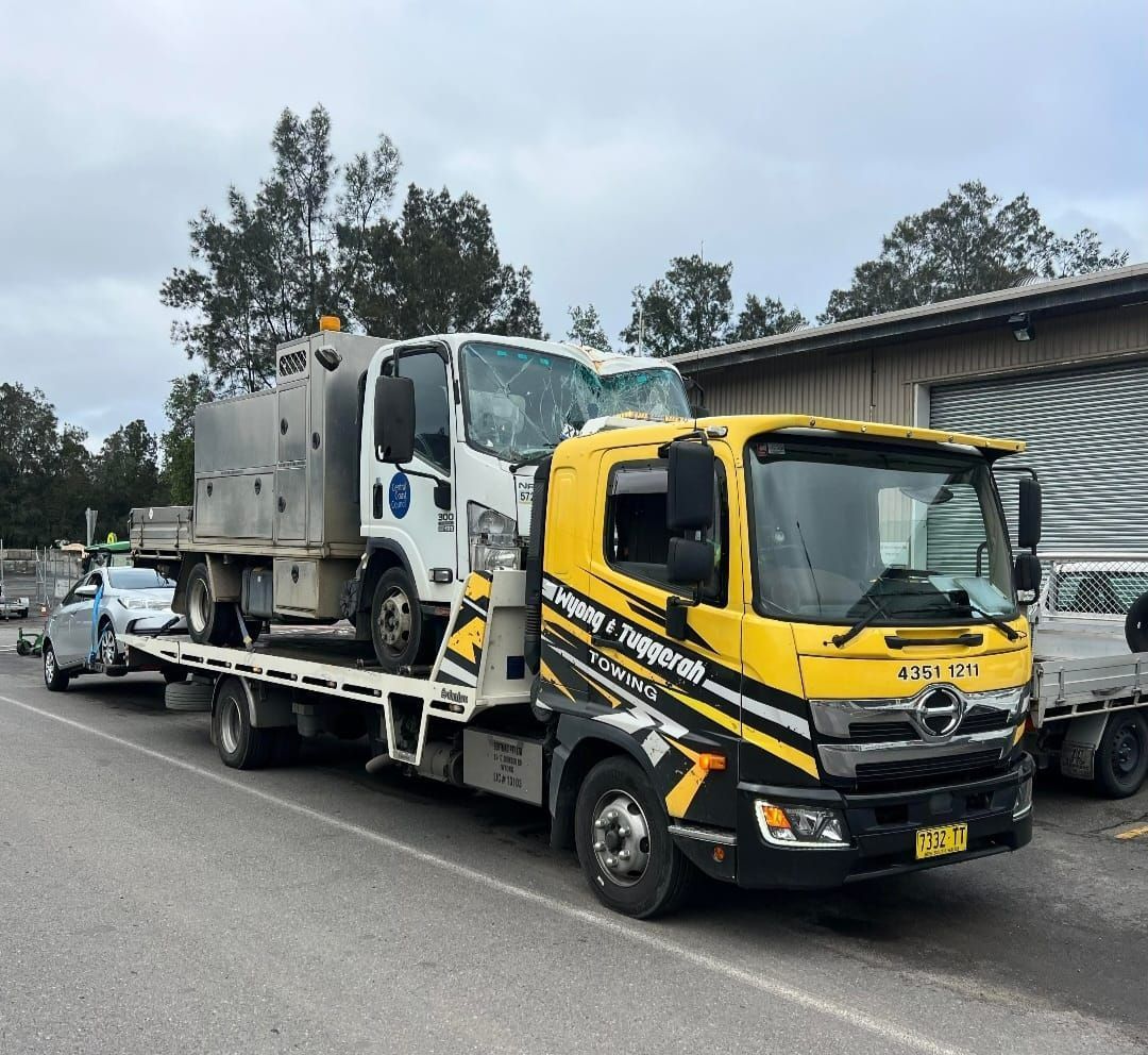 A Yellow Tow Truck is Carrying a Broken Down Truck on a Flatbed Trailer — Wyong and Tuggerah Towing Service in Budgewoi, NSW