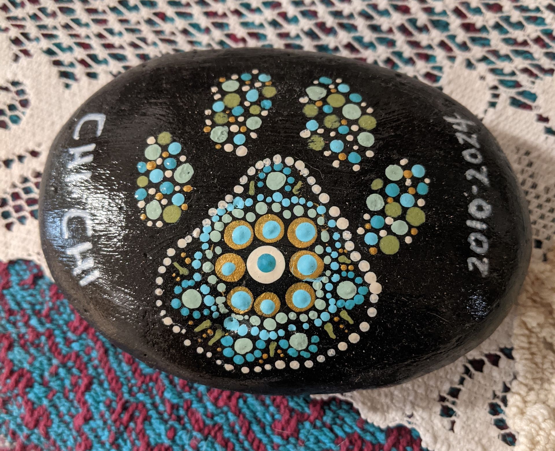 Black painted stone with a turquoise-and-gold paw print and paw shapes on patterned fabric