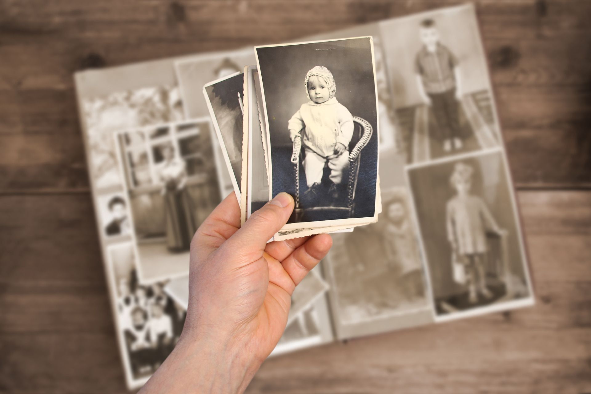 A hand holds a stack of vintage black-and-white portrait photos over an open photo album on a wooden surface.