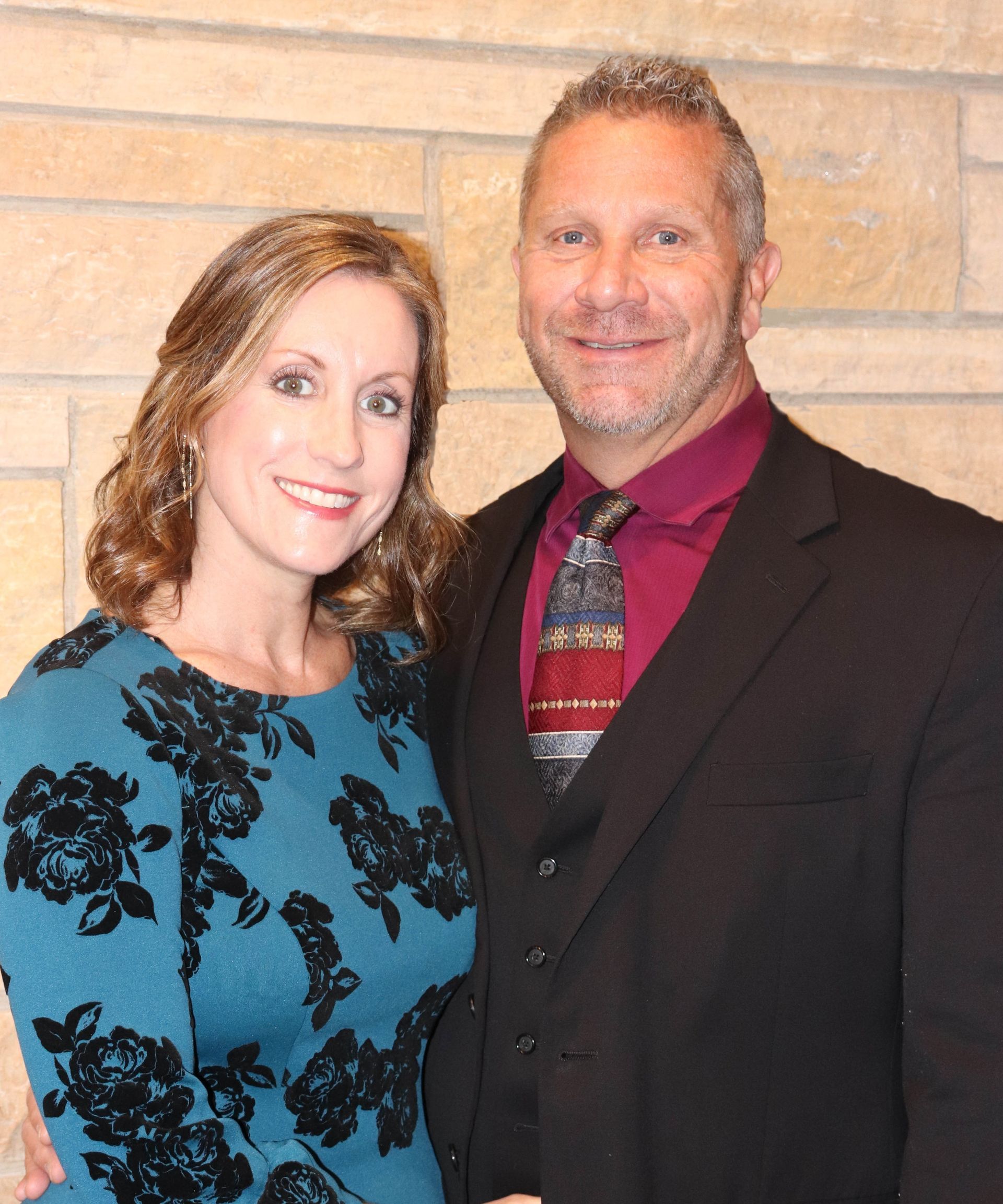A smiling couple poses together against a light-colored brick wall; the woman wears a dark floral top, the man a suit.