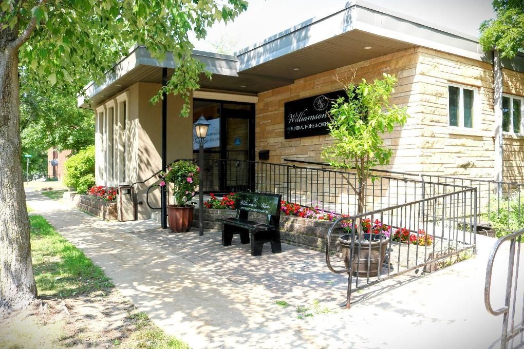 A stone building entrance with a metal railing, ramp, a black bench, and potted flowers under a flat roof.
