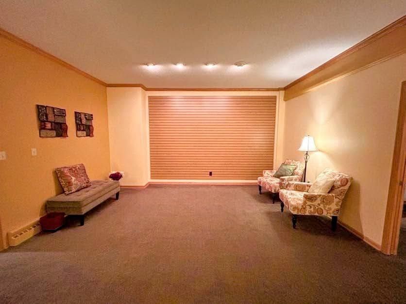 A warm-toned room with carpeted floors, a long bench, two patterned armchairs, and a large horizontal-slatted window.