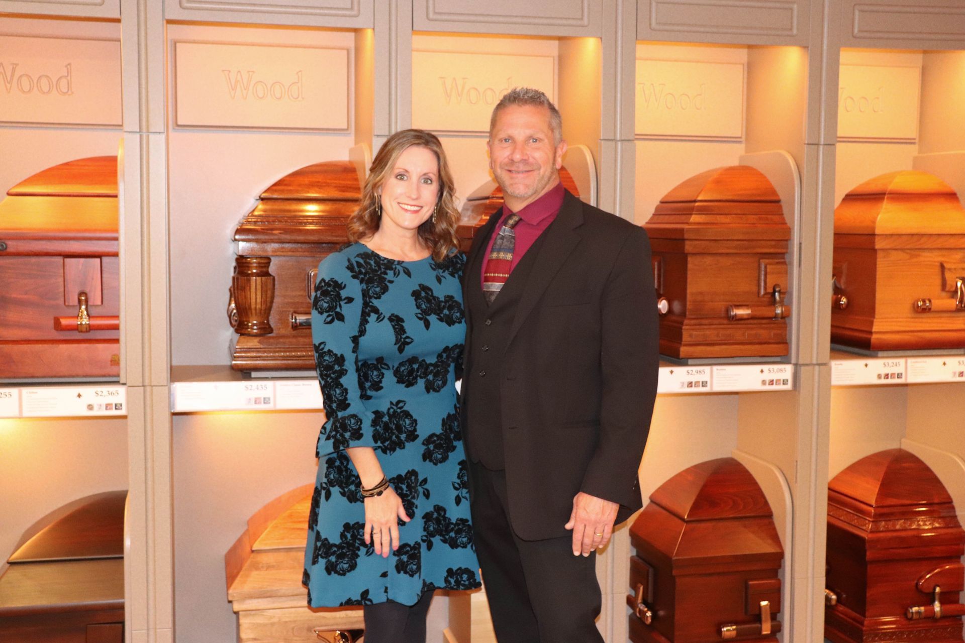 A couple poses smiling in front of a display of wooden caskets in a funeral home showroom.