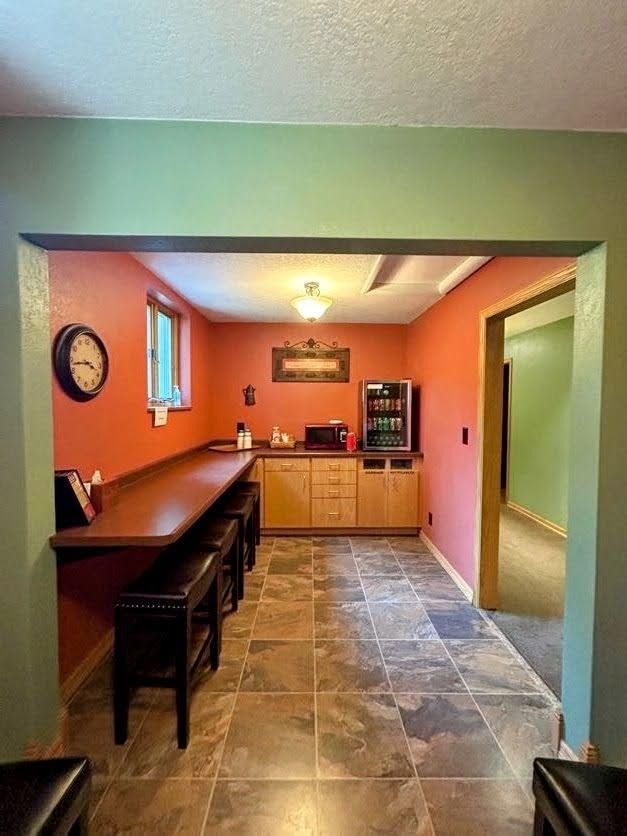 A small kitchen area with an orange breakfast bar, wooden cabinets, a beverage cooler, and stone tile floors.