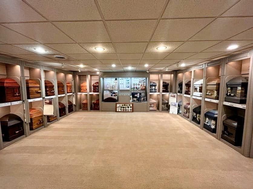 A funeral home showroom featuring shelves displaying various casket styles along three walls under recessed lighting.