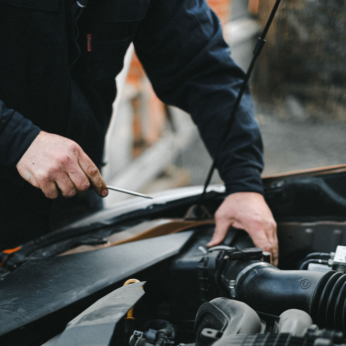 A mechanic in a navy uniform checks a vehicle's engine oil with a dipstick under the open hoo | Ocello's Automotive Center