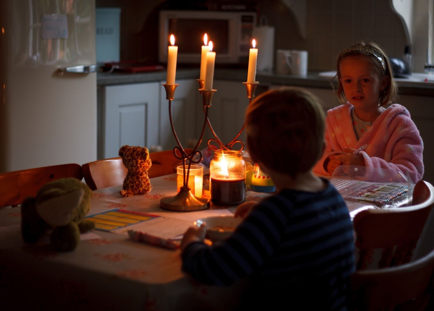 Twee kinderen zitten aan een keukentafel in een schemerige kamer, verlicht door kaarslicht.