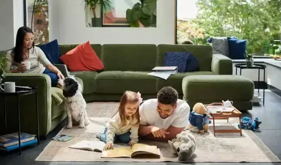 Een gezin en hun hond brengen tijd door in een woonkamer met een hoekbank, waar ze boeken lezen op een vloerkleed.