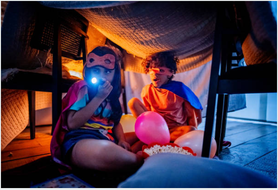 Twee kinderen, als superhelden met capes en maskers, spelen met een zaklamp met ballonnen en een kom popcorn.