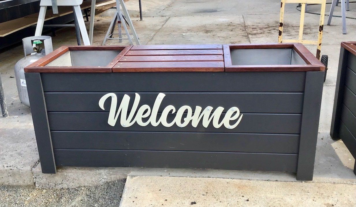 Dark gray wooden planter box with white “Welcome” script on the front outdoors