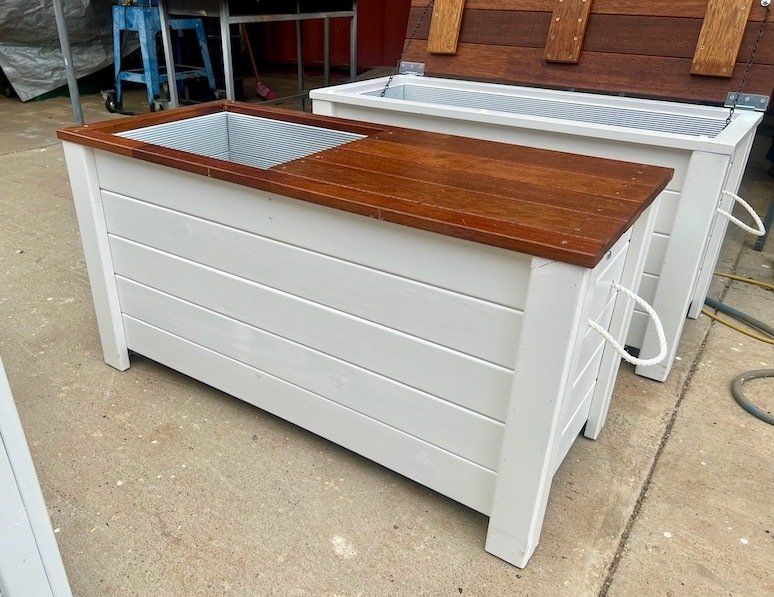 White wooden raised garden beds with brown tops, one with a metal sink insert, on a concrete patio