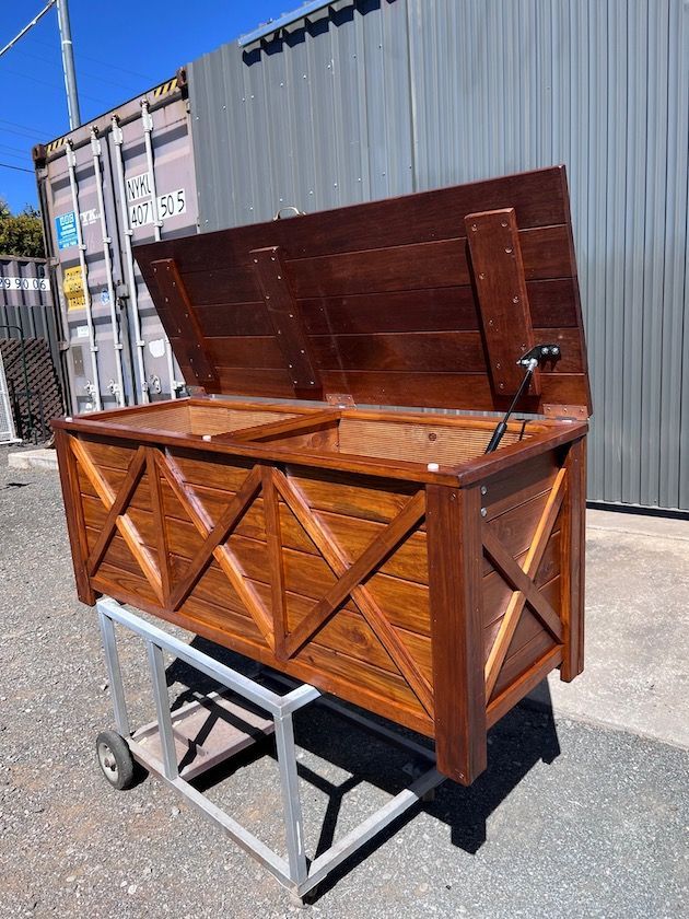 Wooden storage chest on a metal stand outdoors, with the lid open against a corrugated wall.
