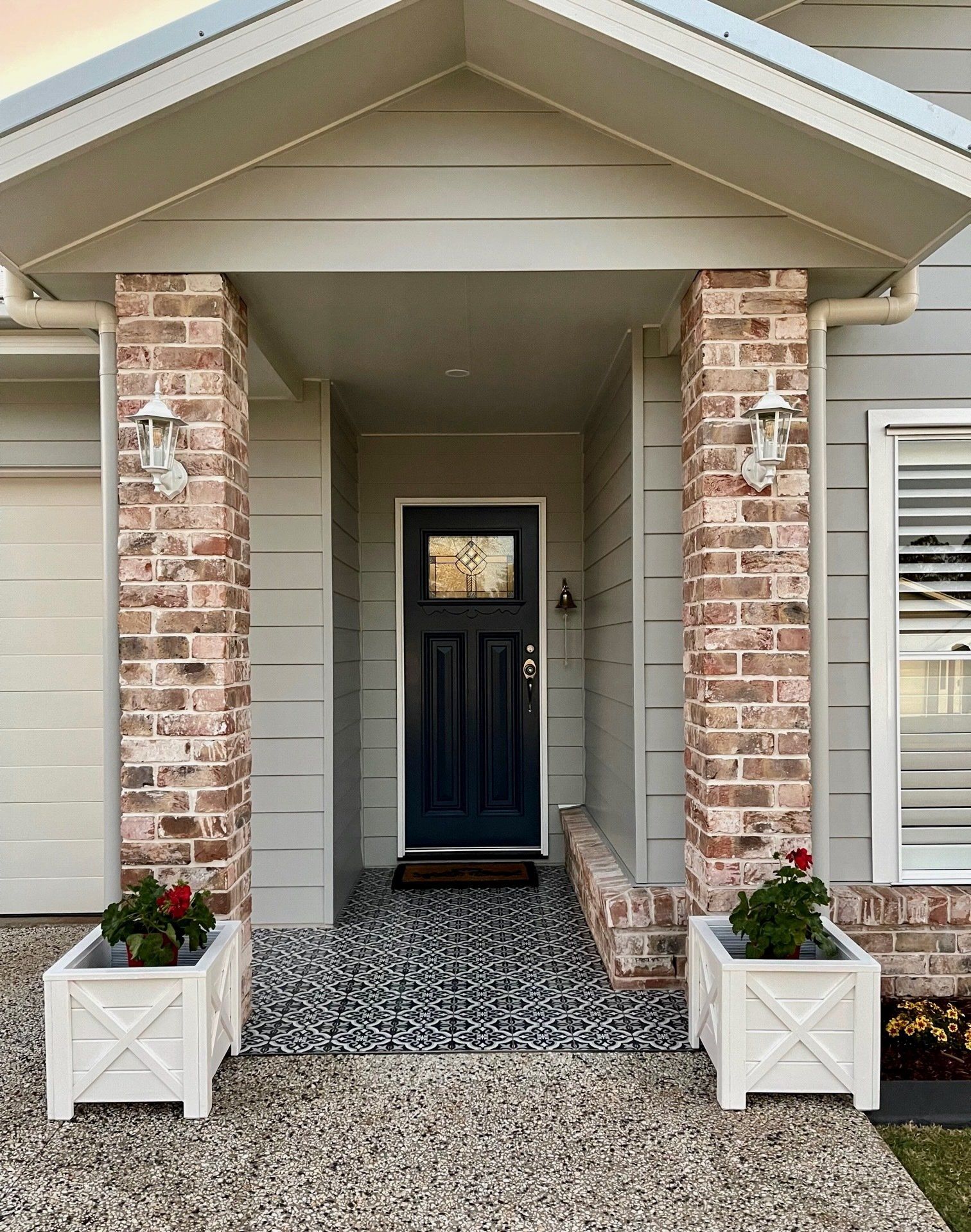 Front porch with brick columns, dark blue front door, and white planters beside a gravel walkway.