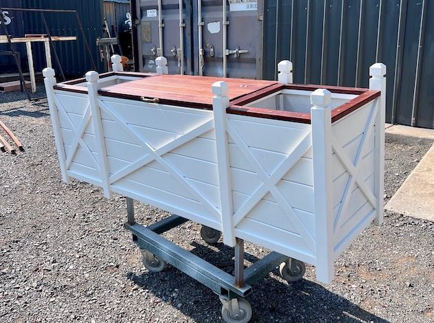 White utility cart on wheels with wooden top, parked on gravel outdoors