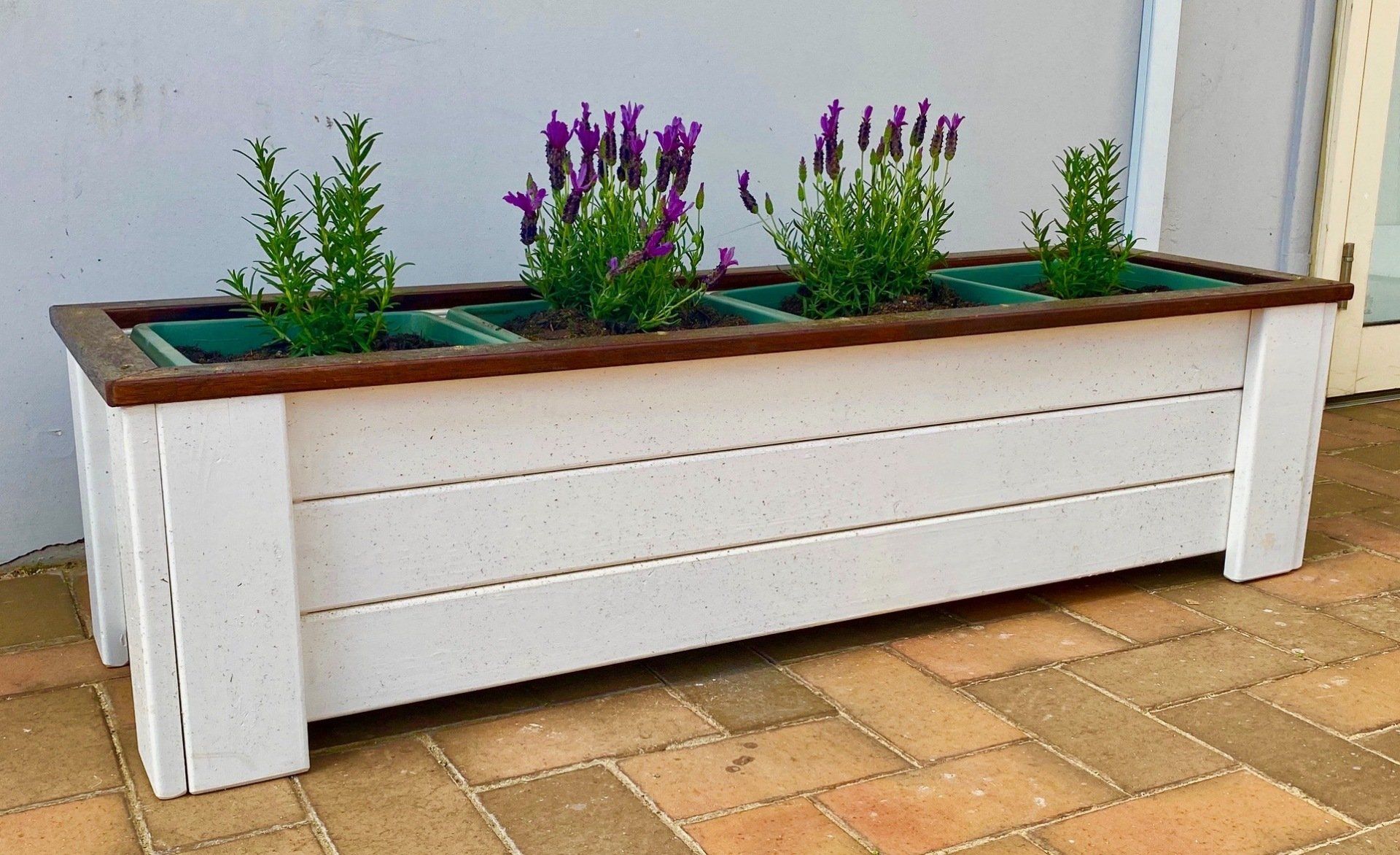 White wooden planter box with purple flowers along a tiled patio wall