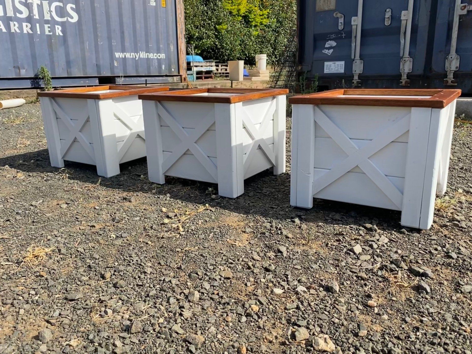 Three white outdoor planter boxes with wooden tops on gravel beside shipping containers.
