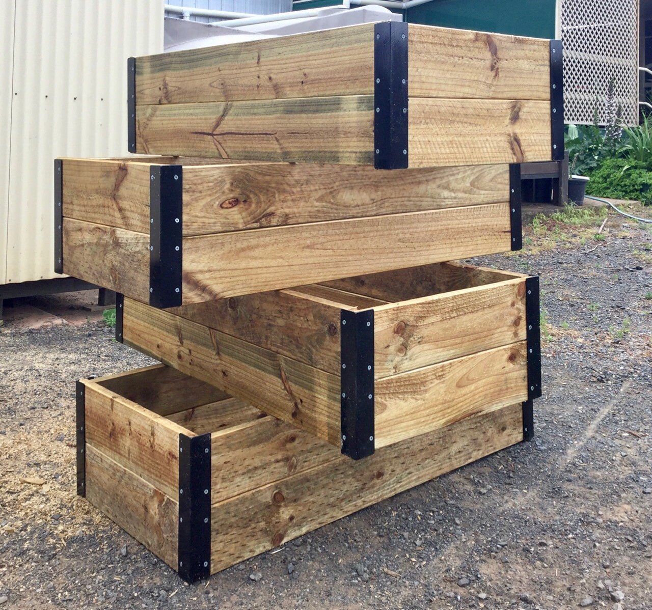 Stacked wooden planter boxes outdoors on gravel, with black metal corner brackets.