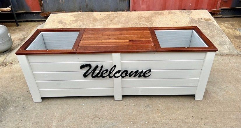 White wooden planter box with wood top, glass corner panels, and black “Welcome” text on the front.