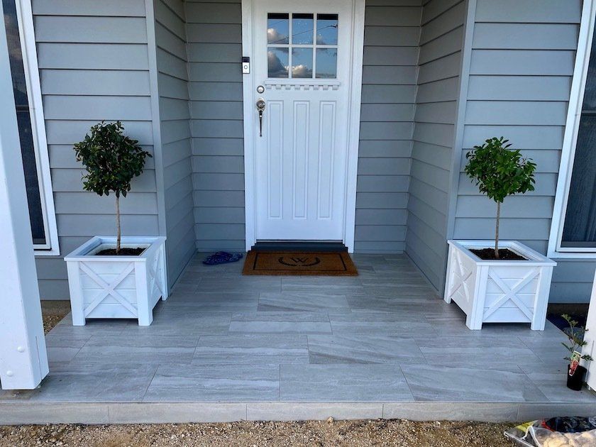 Front porch with white door, gray siding, and two potted topiary trees in white planters.