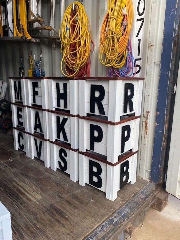 Stacked white lettered display blocks in a workshop, with coiled cables hanging on the wall.