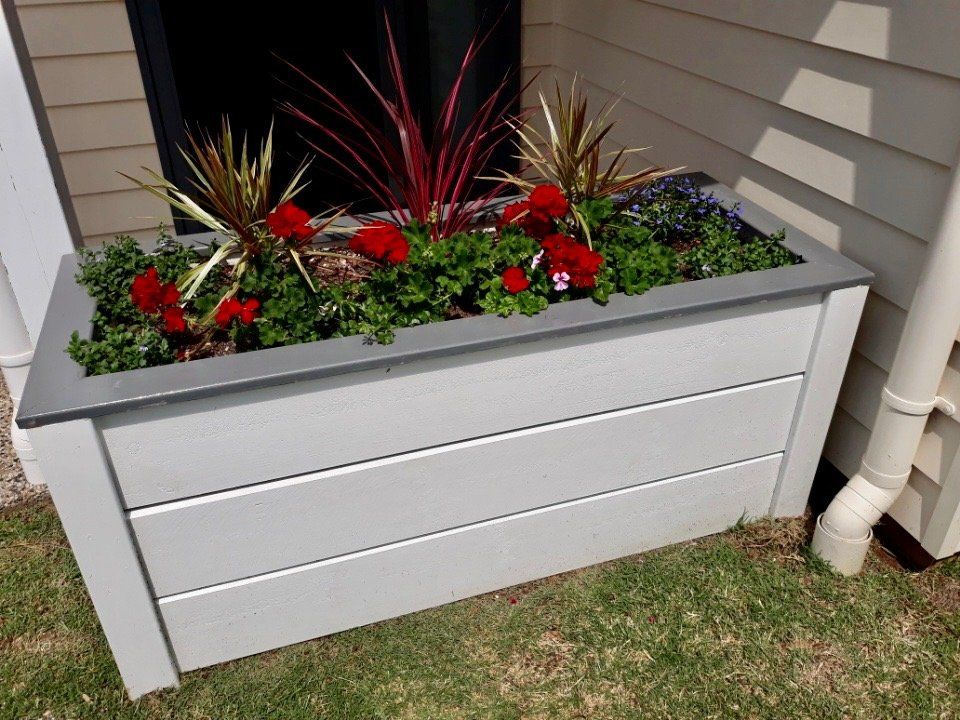 White wooden planter box with red and green flowers beside a house wall and downspout