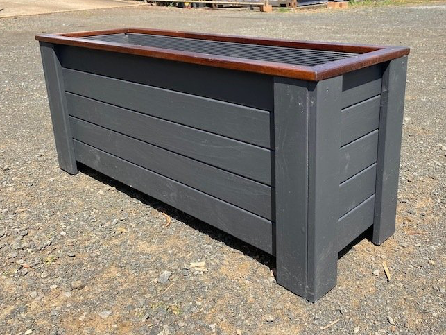 Gray wooden planter box with a brown top trim, shown outdoors on gravel.