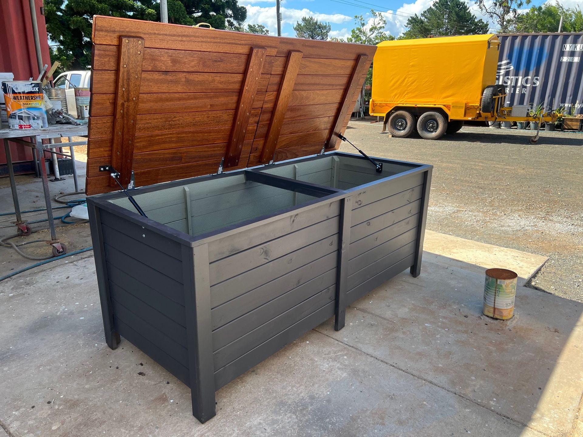 Gray metal raised garden bed with open wooden lid outdoors on a concrete patio.