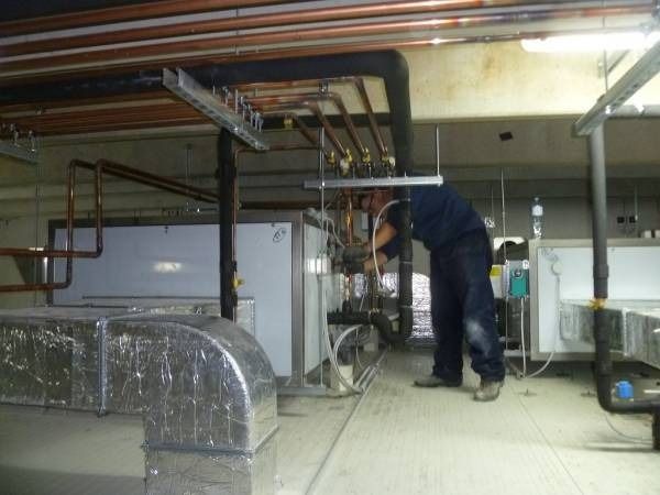 Uomo che lavora sui tubi in una sala macchine, circondato da condotti, tubi di rame e macchinari.