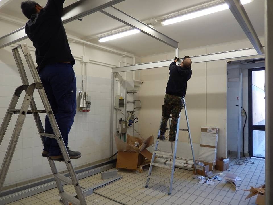 Due operai, in piedi su scale, installano una struttura metallica sul soffitto di una stanza con pavimento piastrellato.
