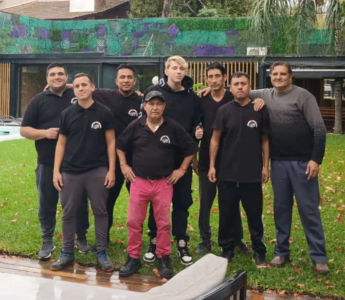 Persona con traje oscuro y corbata a rayas apoyada contra un coche negro en el exterior.