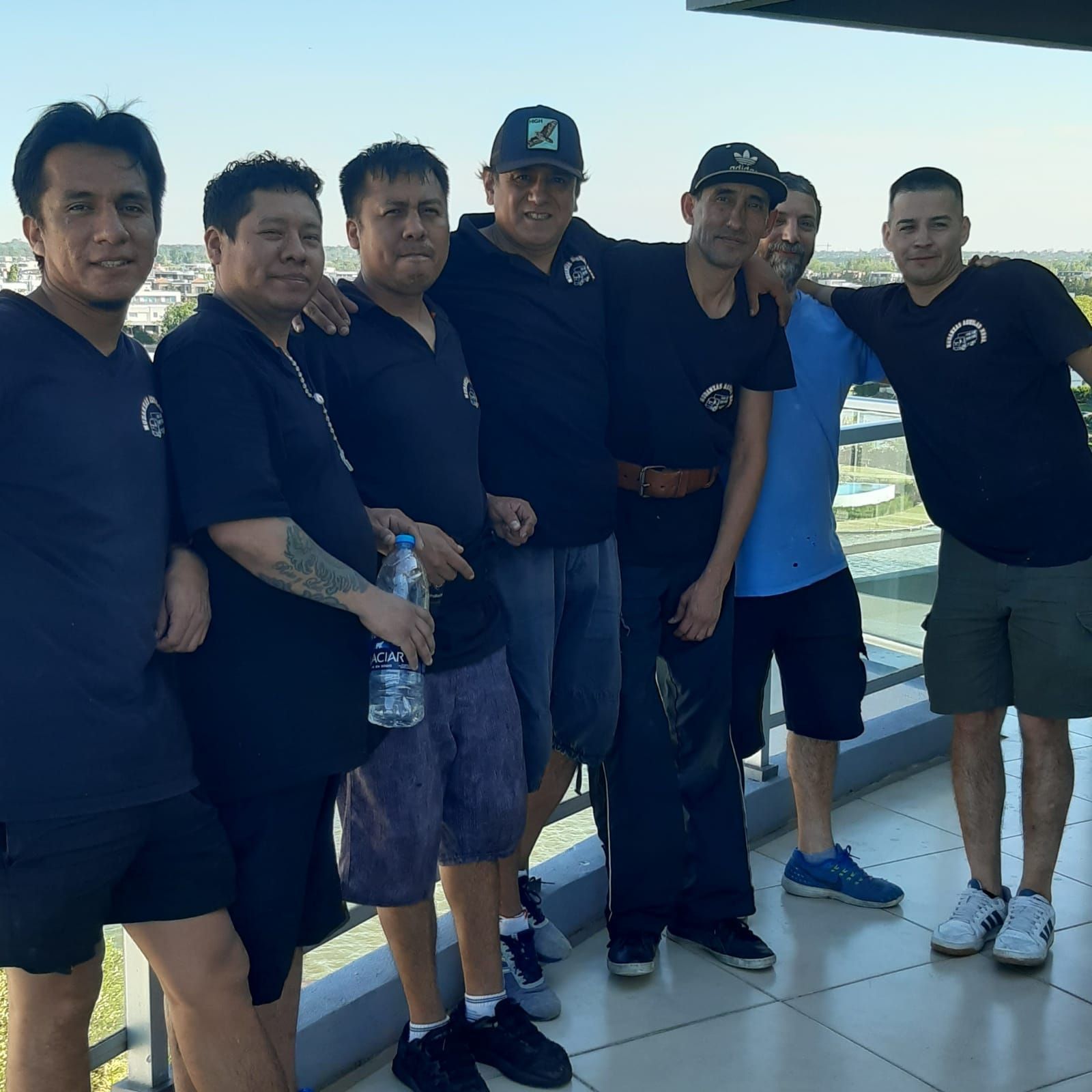 Seis hombres con camisas oscuras posan juntos en una soleada terraza junto al mar; uno de ellos sostiene una botella de agua.