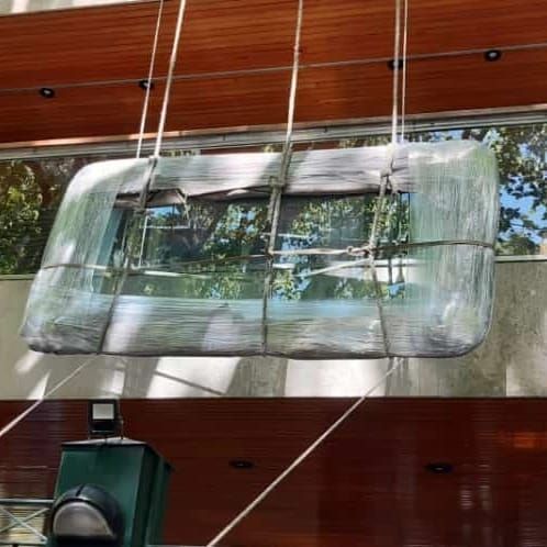 Gran ventanal rectangular de cristal con reflejos, suspendido por cables contra la fachada de un edificio revestida de madera.