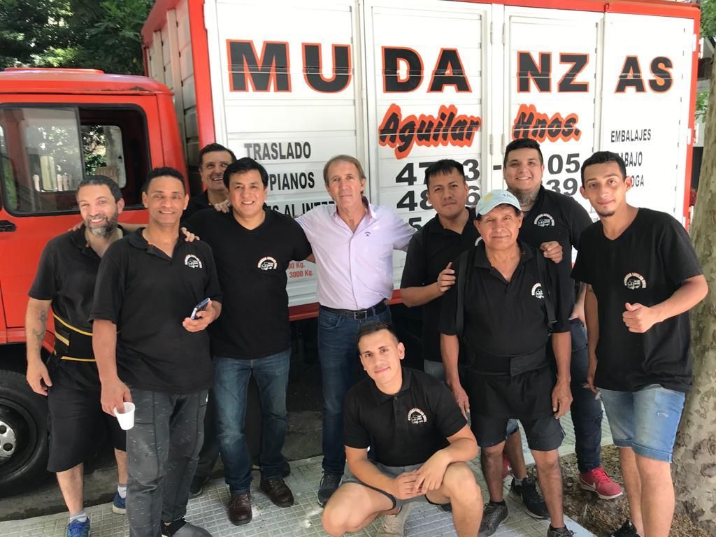 Grupo de personas posando frente a un camión rojo con un letrero de la empresa de mudanzas “Mudanzas”.
