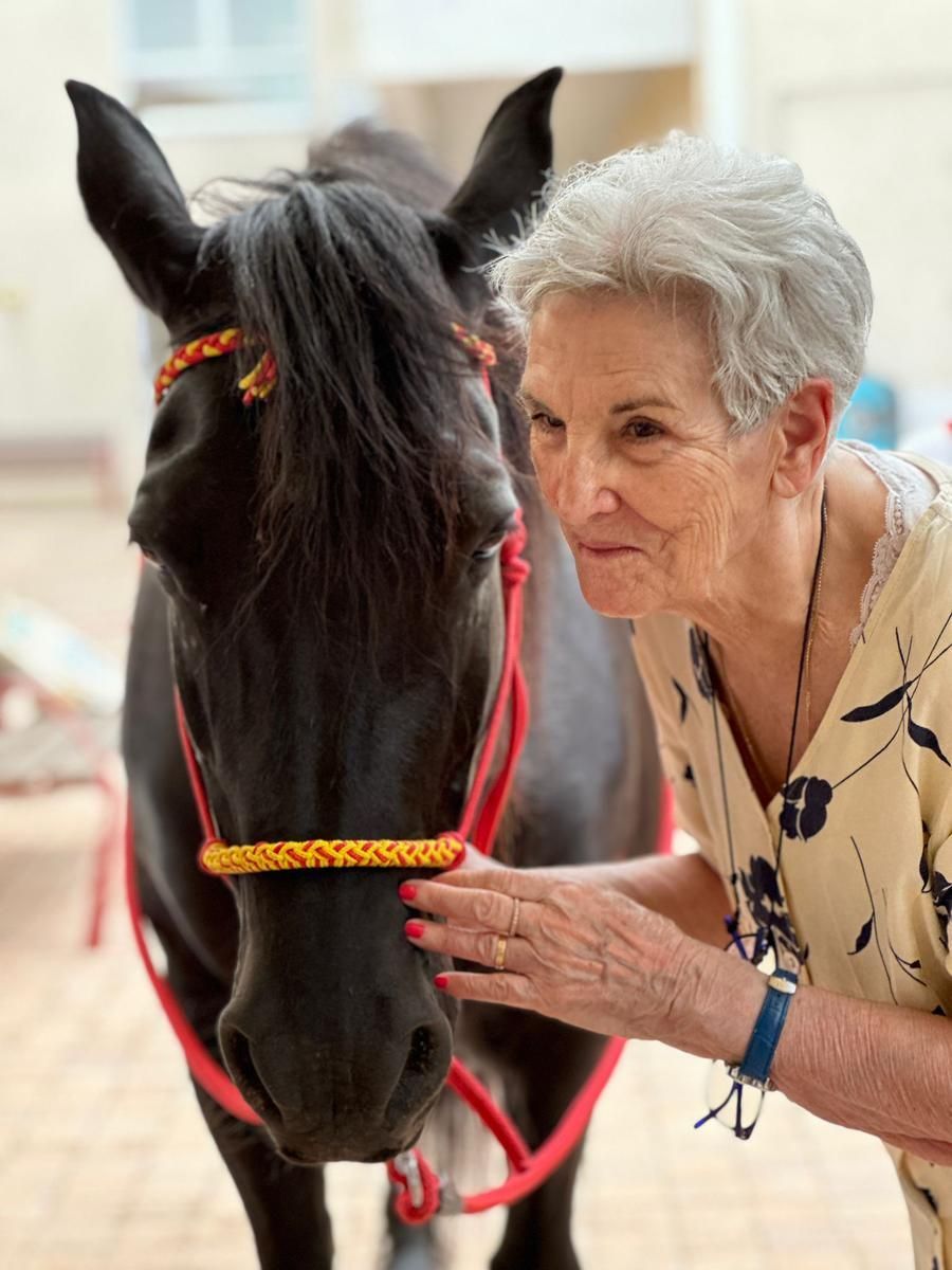 Une femme âgée caresse doucement le museau d'un cheval noir ; ils se trouvent à l'extérieur, devant un bâtiment.