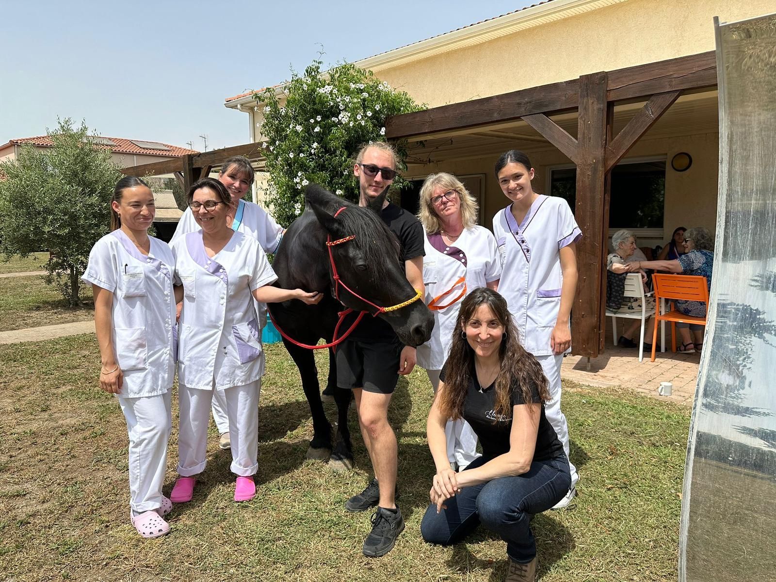 Un groupe de personnes avec un cheval noir à l'extérieur d'un établissement accueillant des résidents, certains souriants.
