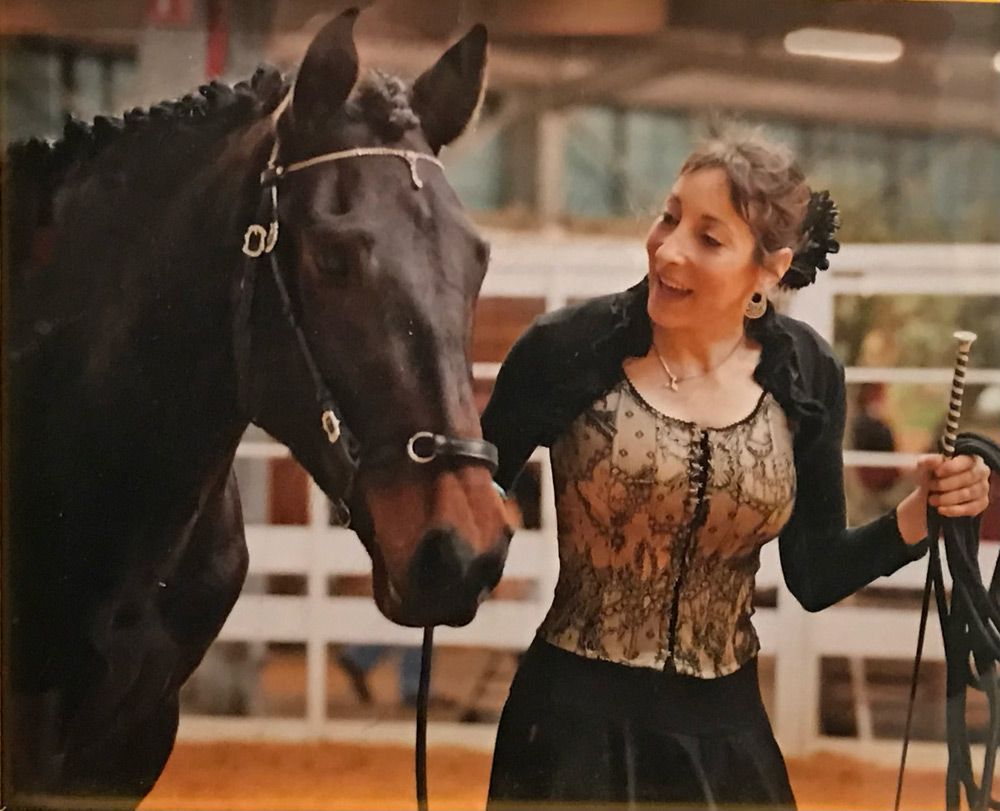 Une femme en tenue de soirée se trouve à l'intérieur, avec un cheval noir. Elle sourit, tenant un fouet ; le cheval porte un bridon.