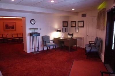 An office interior featuring a wooden desk, blue chairs, a wall clock, framed documents, and red carpeting.