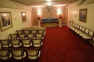 A formal funeral viewing room featuring a casket on a raised stand, draped in blue, with rows of chairs on red carpet.