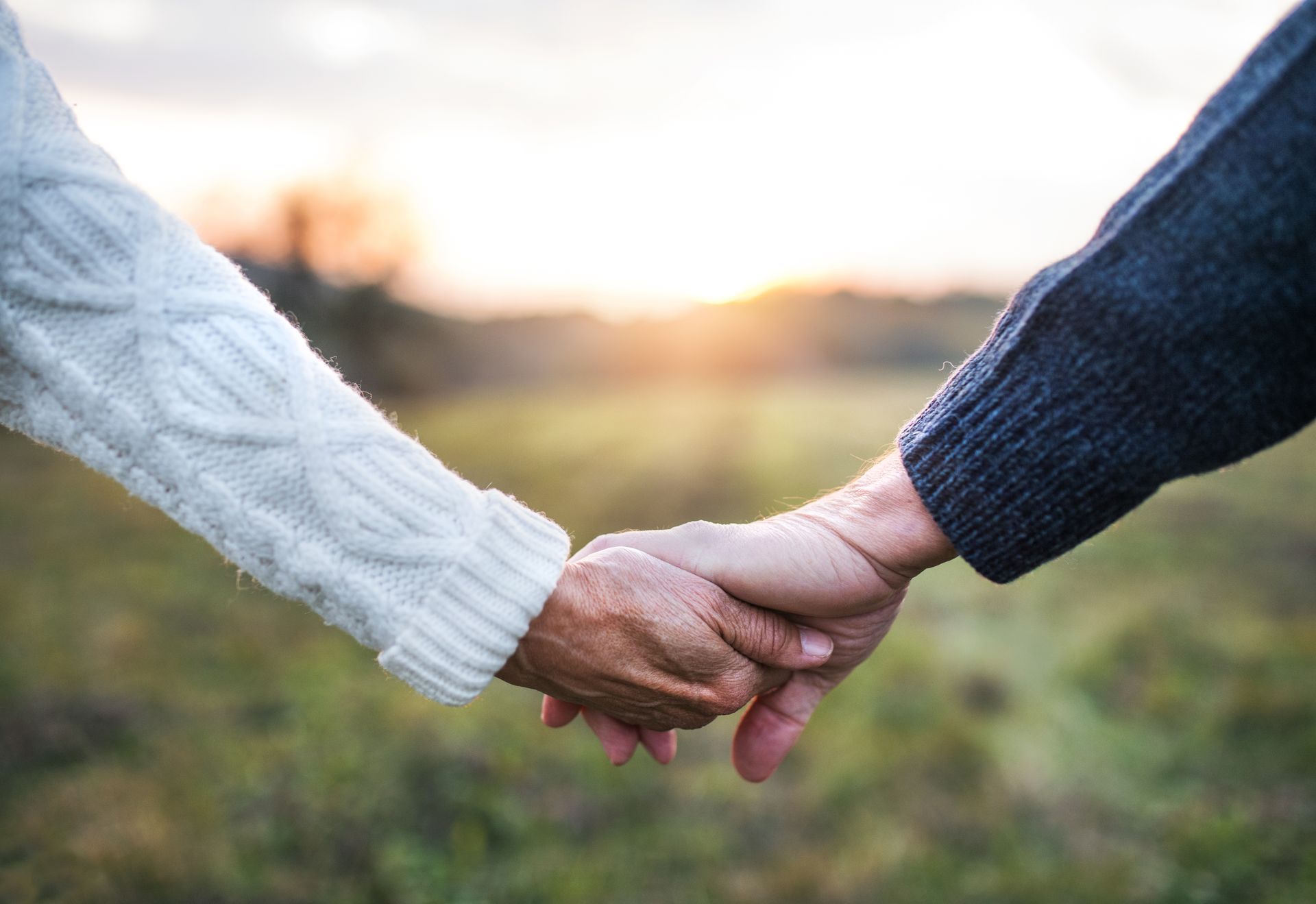Hands clasped, sunset over a field. One person wears a white sweater, the other a navy sweater.