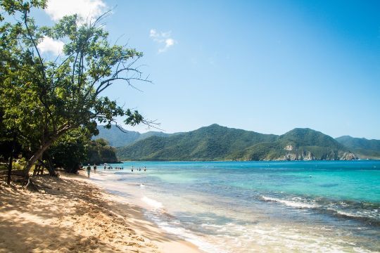 Playa tropical con aguas turquesas, orilla arenosa, árboles y colinas verdes a lo lejos bajo un cielo azul claro.