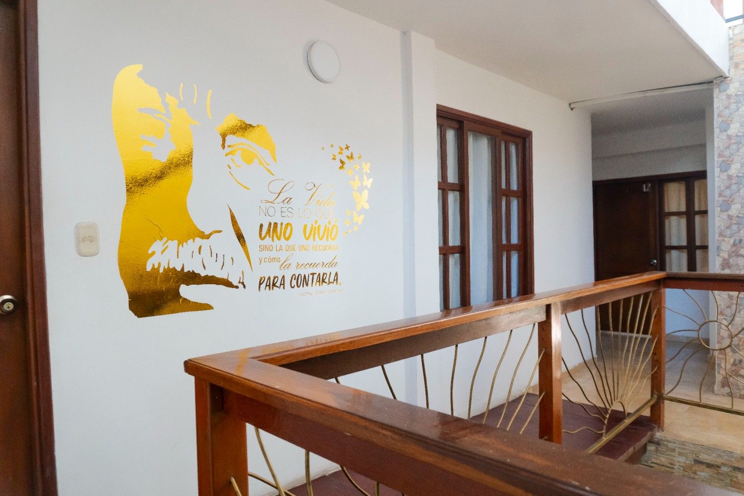 Pasillo interior con paredes blancas, barandilla de madera y un mural amarillo en la pared.