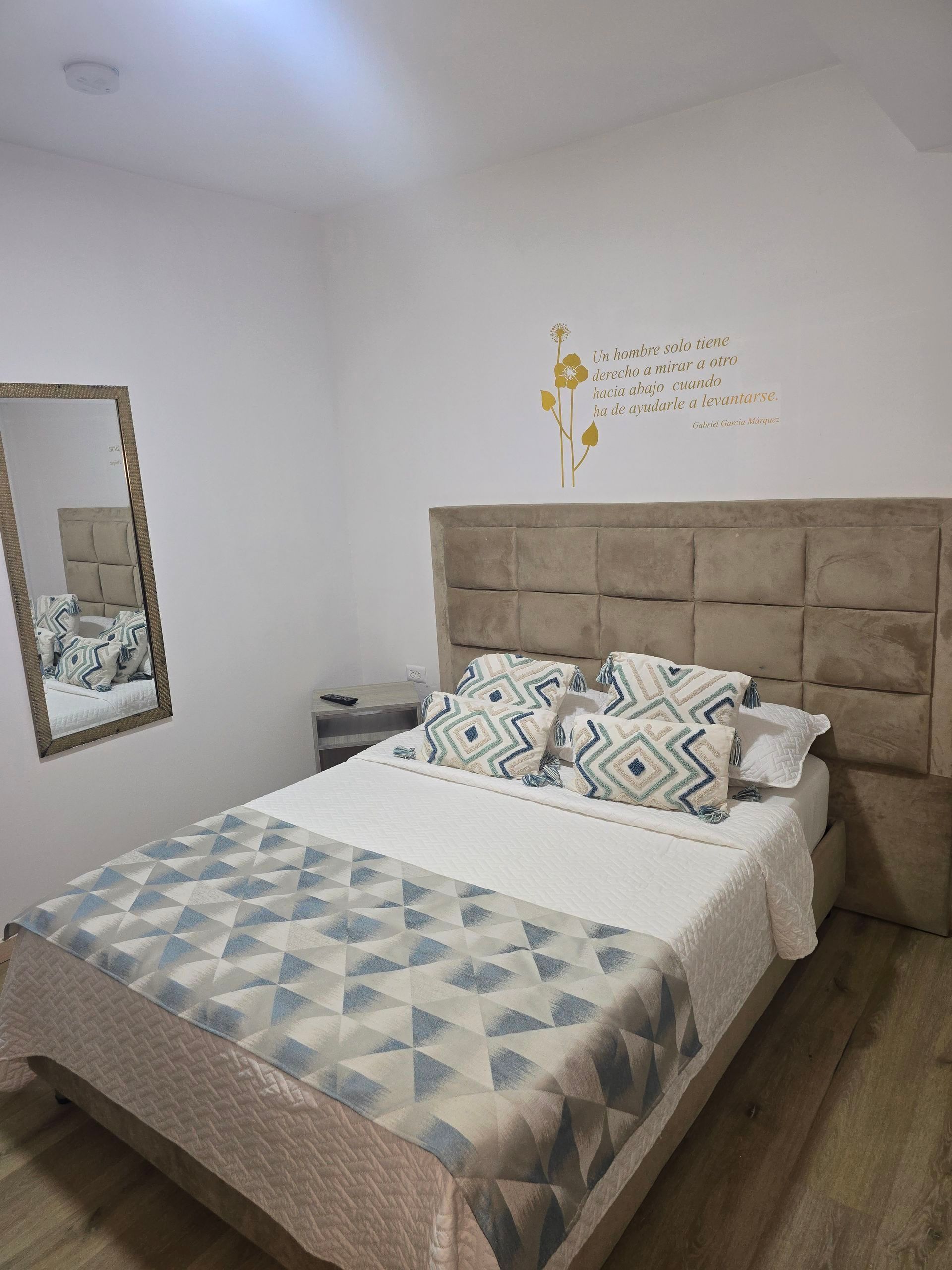 Acogedora habitación con cama tapizada en color beige, ropa de cama estampada, espejo de pared y paredes blancas.