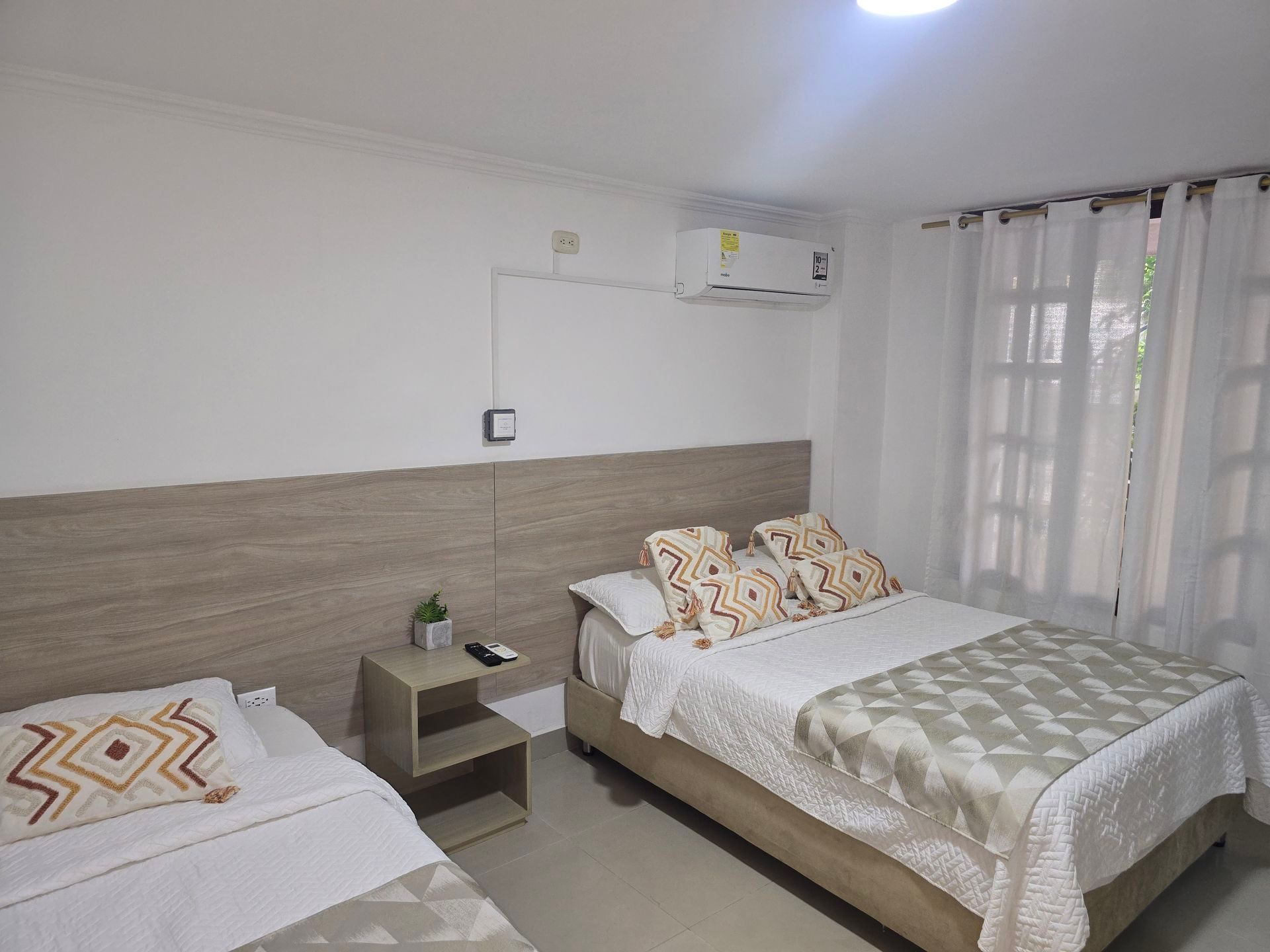 Habitación pequeña con dos camas, revestimiento de madera clara, mesita de noche, aire acondicionado y cortinas transparentes.