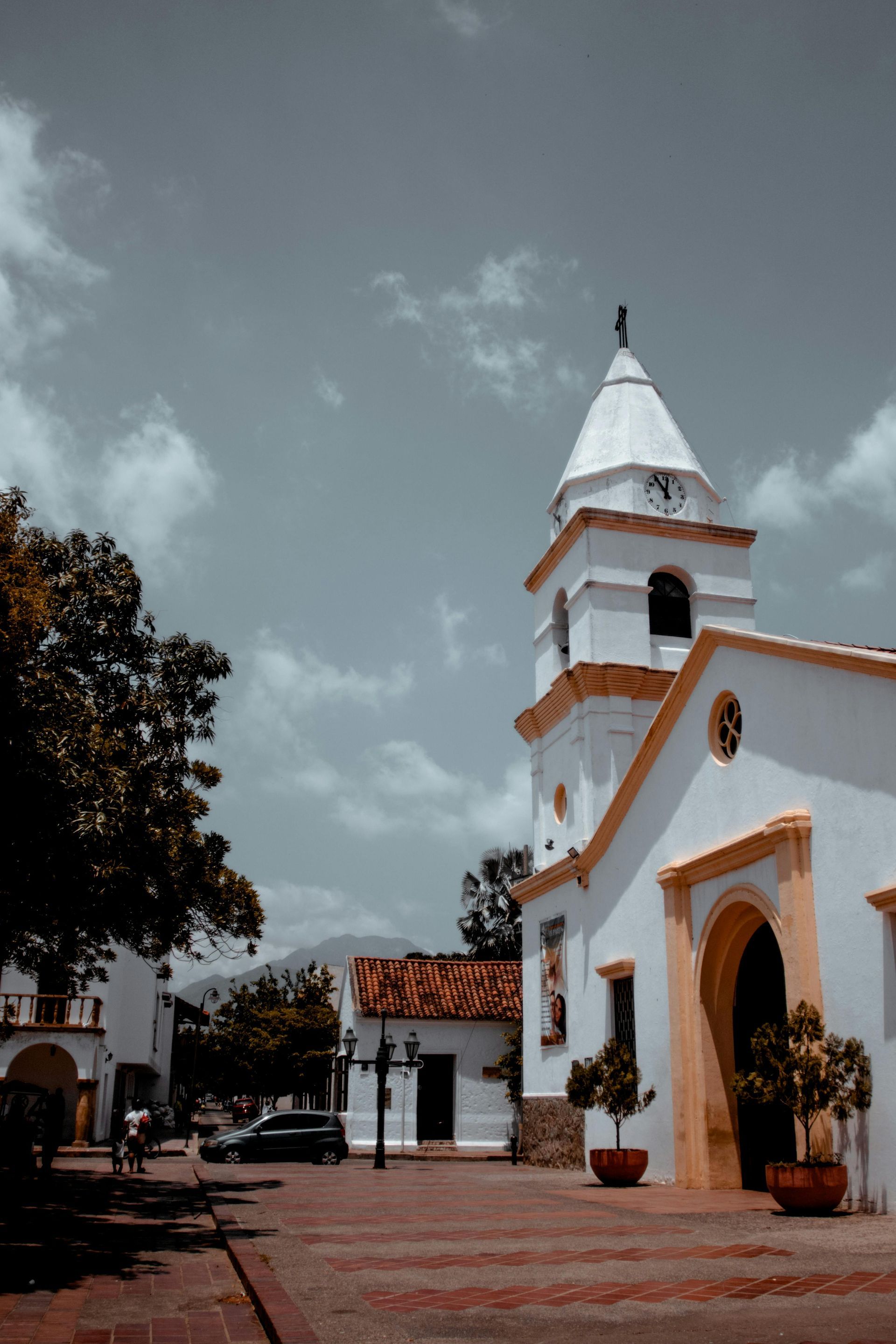 Iglesia blanca con campanario y plantas en macetas en una plaza soleada bajo un cielo nublado.