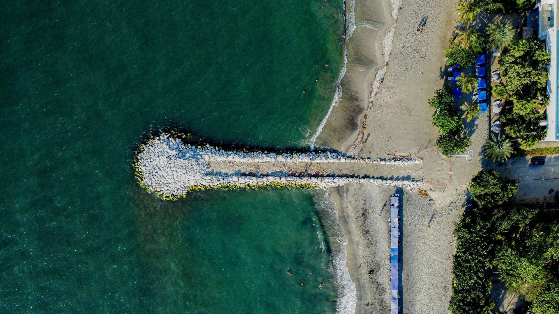 Vista aérea de un muelle rocoso que se adentra en aguas verdes junto a una orilla arenosa y árboles.