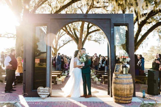 A bride and groom are kissing during their wedding ceremony under a wooden archway.