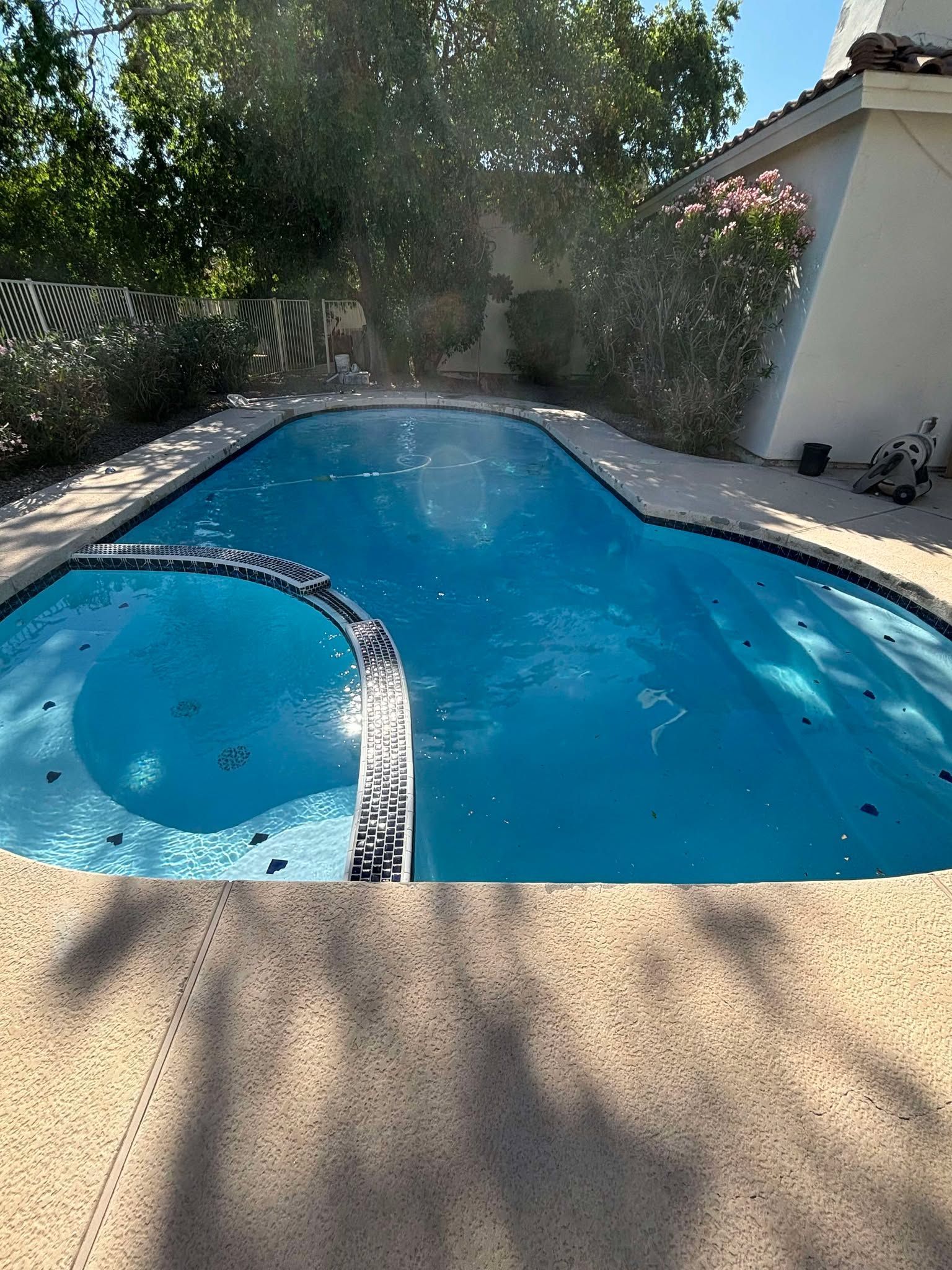 A blue backyard pool with an integrated spa section, surrounded by a light-colored patio and landscaping under a sunny sky.