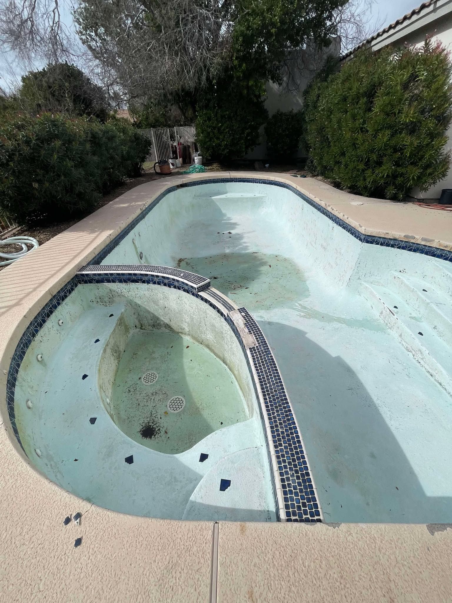 An empty residential swimming pool and attached hot tub with blue tile accents, surrounded by a concrete patio and bushes.