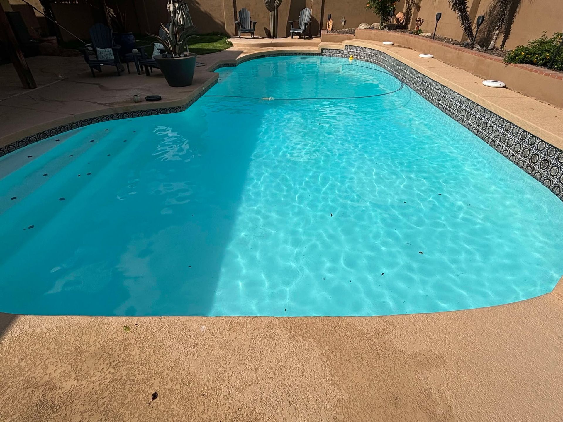 A kidney-shaped swimming pool with blue water and stepped entry, surrounded by a tan deck in a residential backyard.