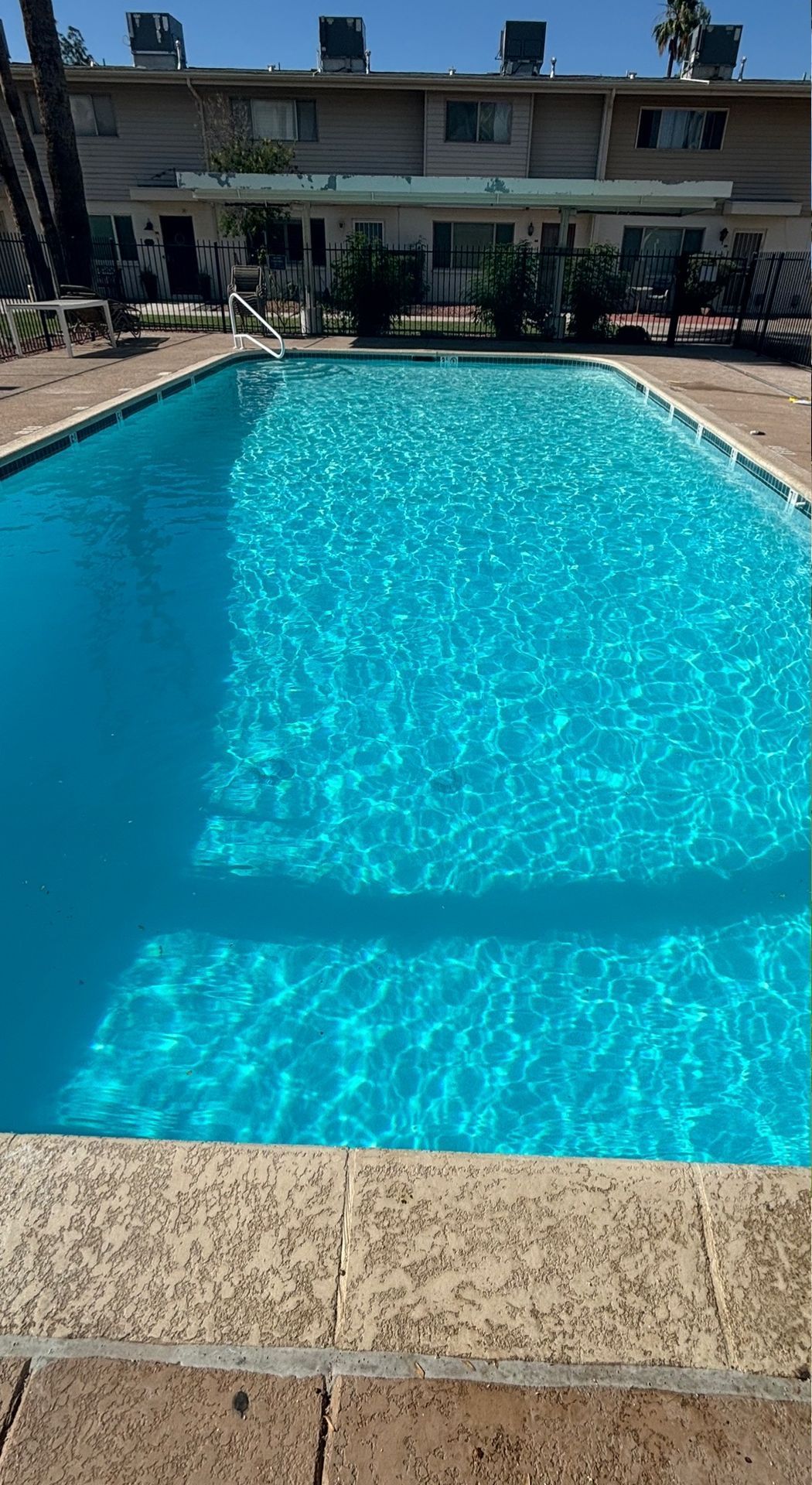 A rectangular swimming pool with bright blue water, surrounded by a tan stone deck in front of a multi-story apartment.
